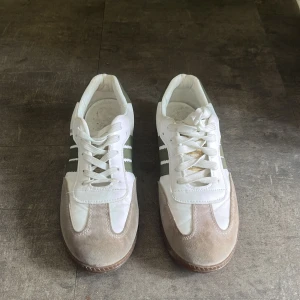 Adidas Samba sneakers i vitt och grönt - Säljer ett par Adidas Samba sneakers i vitt med gröna detaljer och beige mocka framtill. Skorna har klassiska tre ränder på sidan, snörning och gummisula. Perfekta för en sportig och trendig look.