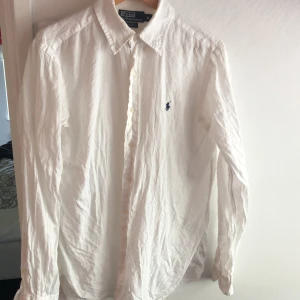 Vit linneskjorta från Polo Ralph Lauren - Klassisk vit skjorta från Polo Ralph Lauren i linne med broderad blå logga på bröstet. Skjortan har lång ärm, knappar framtill och en tidlös krage. Perfekt för en stilren och avslappnad look. 