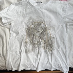 Vit t-shirt med abstrakt tryck - Säljer en vit t-shirt med ett abstrakt, konstnärligt tryck i grått och beige på framsidan. T-shirten har rund halsringning och korta ärmar. Perfekt för en avslappnad och stilren look.