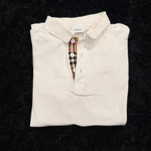 Vit pikétröja från Burberry - Säljer en klassisk vit pikétröja från Burberry med korta ärmar och snyggt rutigt tyg vid knäppningen. Tröjan har broderad logga på bröstet och krage. Perfekt för en stilren look.