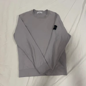 Ljusgrå sweatshirt från Stone Island - Säljer en stilren ljusgrå sweatshirt från Stone Island med klassisk logotyp-patch på ärmen. Tröjan har rund halsringning och långa ärmar. Perfekt för en avslappnad och trendig look.