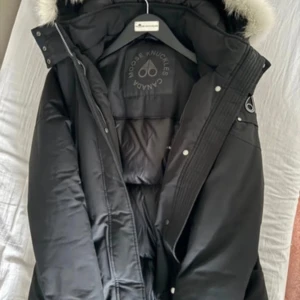 Svart vinterjacka med päls från Moose Knuckles - Säljer en svart vinterjacka från Moose Knuckles med vit päls på huvan. Jackan har lång ärm, dragkedja och knappar framtill samt logotyp på insidan. Perfekt för kalla dagar under vintertid 