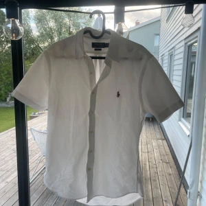 Vit kortärmad skjorta från Polo Ralph Lauren - Klassisk vit skjorta från Polo Ralph Lauren med korta ärmar och knappar framtill. Skjortan har en broderad logga på bröstet och är i storlek M. Perfekt för en stilren och fräsch look. Nice att ha en skjorta som inte är långärmad till sommaren