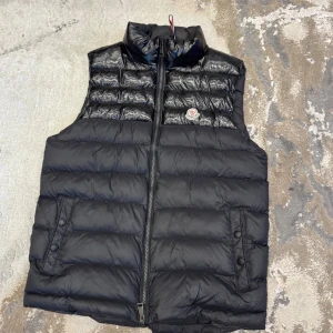 Svart dunväst från Moncler - Säljer en svart dunväst från Moncler med hög krage och dragkedja framtill. Västen har två fickor med knappar och klassisk Moncler-logga på bröstet. Insidan är fodrad i rött tyg och har innerfickor. !!obs!! Västen är inte äkta, och är enda saken på min profil som inte är äkta