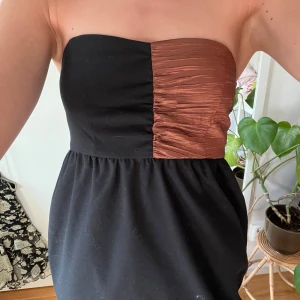 Svart och koppar axelbandslös klänning - Säljer en snygg axelbandslös klänning i svart med en kopparfärgad, glansig detalj upptill. 