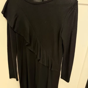 Svart klänning med volang  - Säljer en svart långärmad klänning med rund halsringning och sned volangdetalj framtill. Klänningen har en enkel och stilren design som passar perfekt till olika tillfällen.