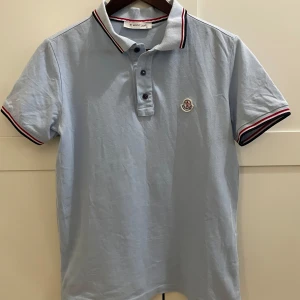 Ljusblå pikétröja från Moncler - Säljer en ljusblå pikétröja från Moncler med klassisk krage och knappar framtill. Tröjan har korta ärmar och detaljer i rött, vitt och blått på krage och ärmslut. Moncler-logga broderad på bröstet. Perfekt för en stilren look.