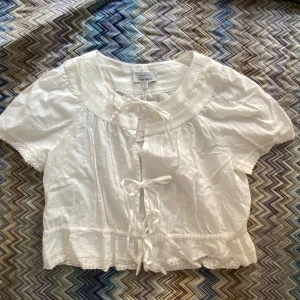 Vit blus med spetsdetaljer från & Other Stories - Supersöt vit blus från & Other Stories med korta ärmar, knytband framtill och fina spetsdetaljer vid krage och ärmslut. Tillverkad i 100% ekologisk bomull och har en romantisk, luftig känsla. Perfekt för vår och sommar! Oandvänd med lappen kvar Orginal pris 590kr. 