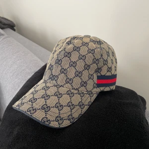 Beige keps från Gucci med monogram - Säljer en beige keps från Gucci med klassiskt GG-monogram i mörkblått och röd-blå rand på sidan. Kepsen har böjd skärm och justerbar passform. Perfekt accessoar för en trendig look.