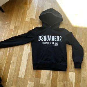 Svart hoodie från Dsquared2 - Säljer en svart hoodie från Dsquared2 med vit text 'DSQUARED2 CERESIO 9, MILANO' på bröstet. Klassisk huva och muddar vid ärmslut och nederkant. Perfekt för dig som gillar streetwear och vill ha något stilrent.