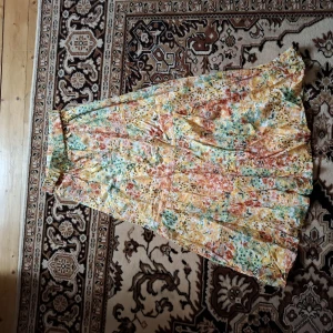 Blommig långkjol i somriga färger - Säljer en långkjol med blommigt mönster i gula, gröna och orange toner. Kjolen har resår i midjan och ett luftigt, flowy fall. Perfekt för varma dagar och ger en bohemisk vibe.