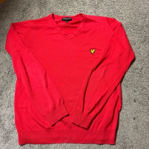 Röd v-ringad tröja från Lyle & Scott - Säljer en röd v-ringad tröja från Lyle & Scott med deras klassiska logga broderad på bröstet. Tröjan har långa ärmar och ribbade muddar vid ärmslut och nederkant. Perfekt för en stilren och enkel look.