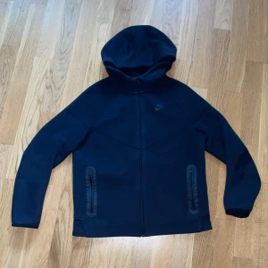 Nike Tech hoodie - Säljer en svart hoodiejacka från Nike med dragkedja framtill och två fickor med dragkedja. Jackan har en stilren design och är perfekt för dig som gillar sportig stil. Passar bra till både träning och vardag.