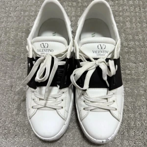 Valentino Garavani | as feta | ev byte - Snygga vita sneakers från Valentino Garavani med bred svart rem över mitten och texten 'OPEN FOR A CHANGE' på sidan. Skorna är i fint skick , kan bytas mot förslag