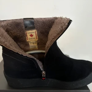 Svarta boots från Canada Snow med dragkedja - Svarta boots i mocka från Canada Snow med mjukt foder och dragkedja på sidan. Skorna har en rund tå och platt sula, perfekta för kalla dagar. Insidan är fodrad med beige, lurvig textil för extra värme.