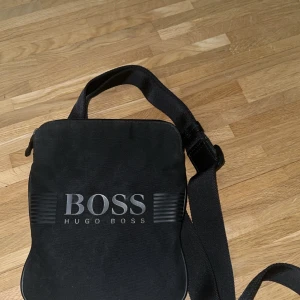 Svart axelväska från Hugo Boss - Snygg svart axelväska från Hugo Boss med justerbar rem och stort logotryck på framsidan. Väskan har dragkedja upptill och är perfekt för att bära med sig det viktigaste. Enkel och stilren design som passar till många tillfällen.