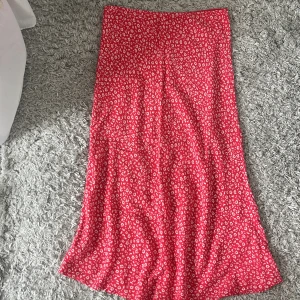 Röd blommig långkjol - Säljer en röd långkjol med små vita blommor. Kjolen har en rak och flowy passform som ger en avslappnad och trendig look. Perfekt att styla med en enkel topp eller hoodie för en cool vardagsstil.