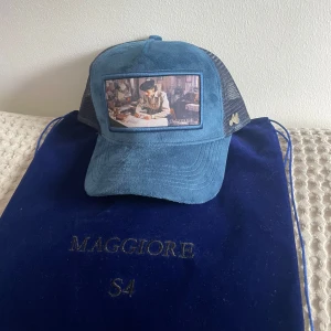 Blå keps från Maggiore med konstmotiv - Snygg blå keps från Maggiore med nät på sidorna och ett broderat konstmotiv framtill. Kepsen har böjd skärm och justerbar passform. Märkets logga i metall på sidan. Perfekt accessoar för en cool och avslappnad stil.