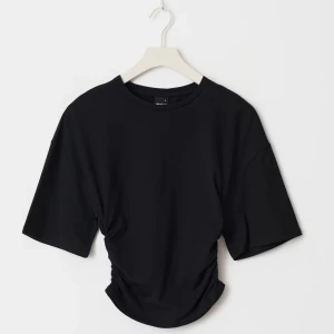 Svart croppad t-shirt med rynkade sidor - Säljer en svart croppad t-shirt med korta ärmar och rynkade detaljer i sidorna. T-shirten har en klassisk rund halsringning och är tillverkad i mjuk bomull. Perfekt för en trendig och avslappnad stil.