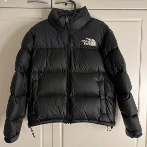 Svart dunjacka från The North Face - Säljer en svart dunjacka från The North Face i storlek M. Jackan har lång ärm, dragkedja framtill och två fickor med dragkedja. Klassisk pufferstil med logga på bröstet och ryggen. 