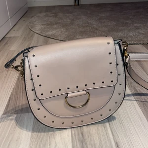 Beige axelväska med nitar och gulddetaljer - Säljer en beige axelväska med rundad form och guldfärgad kedja samt ringdetalj framtill. Väskan har små nitar längs kanterna och ett lock som stängs med magnetknapp. Perfekt för dig som vill ha en stilren men ändå edgy accessoar. Pris kan diskuteras❤️
