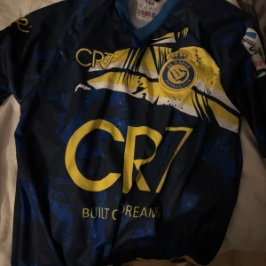 Al Nassr CR7 fotbollströja - Snygg blå fotbollströja från Al Nassr med gult CR7-tryck och Ronaldo 7 på ryggen. Tröjan har korta ärmar, klubbmärke på bröstet och coolt grafiskt mönster i gult och vitt. Perfekt för fans av Cristiano Ronaldo.