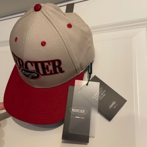 Beige och röd keps från Mercier - Snygg oanvänd keps med lappen kvar från Mercier i beige med röd skärm och röda detaljer. Stor broderad Mercier-logga framtill och texten 'Established 2017' broderad baktill. Justerbar passform och klassisk sexpanel-design.