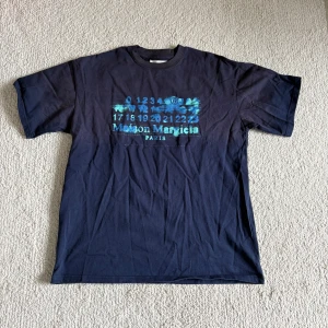 Svart t-shirt från Maison Margiela med blått tryck - Säljer denna blå T-shirten från Maison Margiela! Den är väldigt unik och har en cool design! Perfekt nu till Sommarn!! Storleken är M!( Lite stor i Storleken! Pris: 400! Ej hugget i sten! Hör av dig vid frågor och funderingar!!