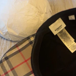 Beige rutig keps från Burberry - Säljer en klassisk keps från Burberry i beige med det ikoniska rutiga mönstret i svart, vitt och rött. Kepsen har böjd skärm och justerbar passform. Perfekt accessoar för en stilren look. pris ej hugget i sten