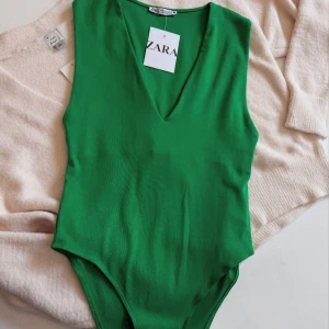 Grön body från Zara - Säljer en snygg grön bodysuit från Zara med v-ringning. Bodyn är ärmlös och har en ribbad struktur som ger en modern look. Perfekt att styla till jeans eller kjol för en trendig outfit.