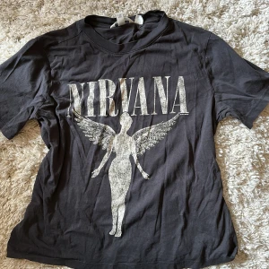 Svart Nirvana t-shirt från H&M - Cool svart t-shirt från H&M med stort Nirvana-tryck och ikonisk ängel på framsidan. T-shirten har rund hals och korta ärmar, perfekt för dig som gillar bandmerch och vill ha en avslappnad stil. Storlek S, oversized.