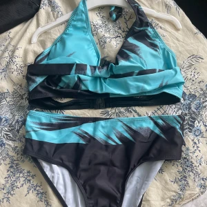 Turkos och svart bikini med grafiskt mönster - Säljer en snygg bikini i turkos och svart med grafiskt mönster. Överdelen har halterneck och knyt i nacken, medan underdelen har normal passform. Perfekt för stranden eller poolen!Helt ny storlek m