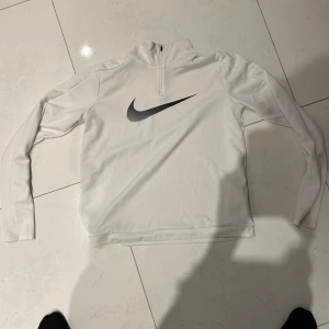 Vit långärmad träningströja från Nike - Säljer en vit långärmad träningströja från Nike med halv dragkedja och stor svart logga på bröstet. Tillverkad i Dri-FIT-material som andas och passar perfekt till träning eller sportaktiviteter.