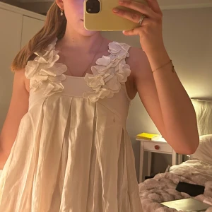 Vit klänning med volangdetaljer - Säljer en söt vit klänning med stora volangblommor vid axlarna. Klänningen har en lös passform och är ärmlös, med veckad kjol som ger ett luftigt intryck. Perfekt för sommarens alla tillfällen!