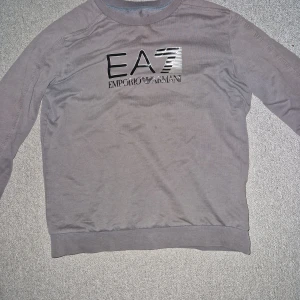 Grå sweatshirt från Emporio Armani EA7 - Säljer en stilren grå sweatshirt från Emporio Armani EA7. Tröjan har rund halsringning, långa ärmar och en stor logga framtill. Perfekt för en avslappnad och trendig look.