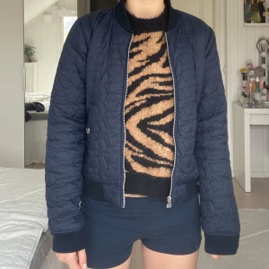 Mörkblå quiltad bomberjacka från Svea - Säljer en mörkblå quiltad bomberjacka från Svea i storlek medium. Jackan har dragkedja framtill, ribbade muddar och två fickor med knappar. Perfekt för dig som vill ha en stilren och sportig look.