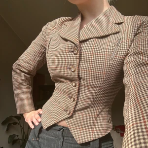 Brun och beige rutig kavaj med markerad midja - Säljer en vintage kavaj i brunt och beige rutigt tyg med bred krage och korsett likande passform. Kavajen är i lite tjockare tyg och är välsydd. Har en liten reva i kragen, se sista bilderna. Perfekt för dig som letar efter en tidsenlig pärla. 