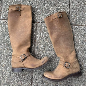 Beiga höga boots med spänne och dragkedja - Säljer ett par höga beiga boots från Primeboots i skinn med snygga spännen och grov dragkedja baktill. Skorna har rund tå och platt sula, perfekt för dig som gillar en klassisk och robust stil. Bra märke likt Frye - Primeboots, RRP är ca 3000kr. 