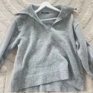 Grå stickad tröja med krage från Gina Tricot - Mysig grå stickad tröja med bred krage och v-ringning från Gina Tricot. Tröjan har långa ärmar och ribbade detaljer vid ärmslut