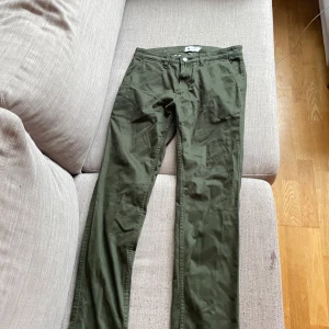 Olivgröna chinos från NN07 - Snygga olivgröna chinos från NN07, tillverkade i italienskt tyg. Klassisk rak passform med dragkedja och knapp framtill samt diskreta bakfickor. Perfekta för en stilren och avslappnad look.