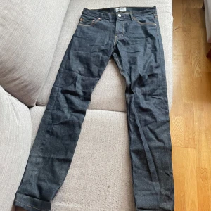 Mörkblå jeansbyxor från WESC - Säljer ett par mörkblå jeans från WESC, modell EDDY. Byxorna har klassisk femficksdesign, raka ben och kontrastsömmar. Snygg detalj med gul patch bak i midjan. Perfekta för dig som gillar stilrena jeans med en twist.
