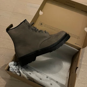 Helt nya Dr. Martens 1460 - Säljer ett par oanvända Dr. Martens 1460 boots i nickelgrå nubuck med svart platt sula och klassisk snörning. Skorna har rund tå och robust design, perfekta för dig som vill ha en tidlös och cool look.