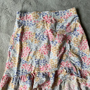 Blommig volangkjol i bomull - Säljer en söt blommig kjol med volangkant. Kjolen har små blommor i rosa, blått och gult på vit botten. Den är kort och har en omlottliknande detalj framtill. Perfekt för vår och sommar!