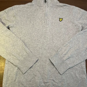Ljusgrå halfzip från Lyle & Scott - Säljer en ljusgrå stickad tröja från Lyle & Scott Junior med halv dragkedja och deras klassiska logga på bröstet. Tröjan har långa ärmar och ribbade muddar. Perfekt för en stilren och bekväm look.