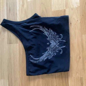 Svart oneshoulder topp med strasstenar - Snygg svart oneshoulder topp med glittrande strasstenar i ett dekorativt mönster framtill. Perfekt för att sticka ut och ge outfiten lite extra edge.