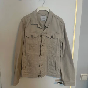 Vårjacka Jack & Jones - Säljer en beige jeansjacka från Jack & Jones med klassisk krage och knäppning framtill. Jackan har två bröstfickor med lock och knappar samt sidofickor. Perfekt för vår. Aldrig använd, prislapp sitter kvar. 