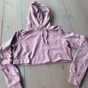 Ljuslila croppad hoodie från Gymshark - Säljer en ljuslila croppad hoodie från Gymshark med huva och långa ärmar. Tröjan har en stilren design med Gymshark-logga på bröstet och ribbade muddar vid ärmsluten. Perfekt för en sportig och avslappnad look.