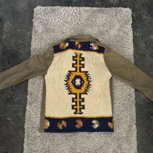 Olivgrön overshirt med mönstrad rygg - Säljer en olivgrön overshirt med fyra stora fickor framtill och knappar. Baksidan har en unik stickad panel med färgglatt geometriskt mönster. Perfekt nu till sommarkvällar osv. Pris går alltid att diskutera vid snabb affär och bytesförslag är intressant! Är 185cm på bilderna!