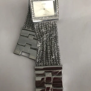 Silverfärgad klocka med glittrande detaljer från Cussi - Säljer en snygg klocka från Cussi med fyrkantig urtavla, glittrande stenar runt ramen och ett bredare armband i silverfärg. Klockan har ett unikt mönster på armbandet och passar perfekt som accessoar.