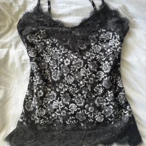 Svart blommig linnetopp med spets från Rosemunde - säljer en svart och grå blommig linnetopp från Rosemunde med tunna axelband och vacker spetskant upptill och nedtill. toppen har ett elegant blommönster och är perfekt för att skapa en stilren look. finns ingen storlek på den, men hade sagt att den passar s-m. aldrig använt, som ny!